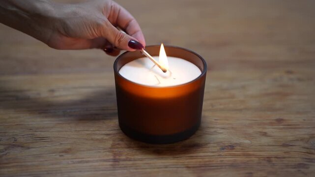 Lighting scented candle with match on wooden table. Cozy home atmosphere, relaxation, aromatherapy and self-care concept.