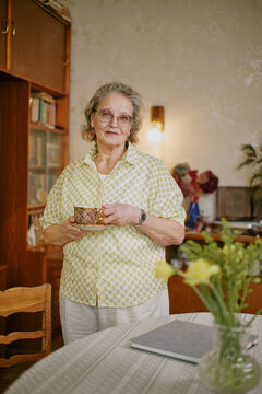 Portrait of senior Caucasian woman standing in home environment holding patterned mug with both hands, wearing glasses and earrings, looking directly at camera with gentle expression