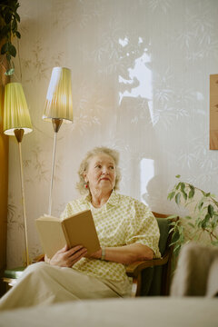 Senior Caucasian woman sitting and holding book, gazing thoughtfully to side, short gray hair and earrings visible, surrounded by indoor plants and standing lamps in living room
