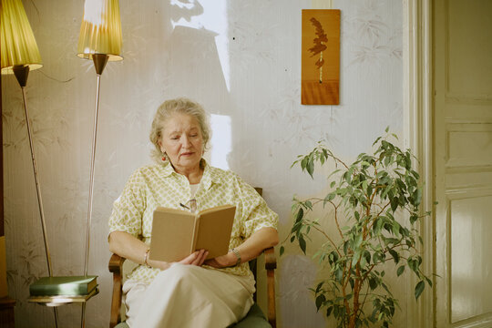 Senior Caucasian woman sitting in armchair reading book, appearing relaxed and focused, indoor setting with houseplant and vintage lamps visible in background