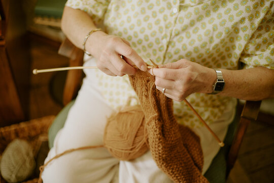 Senior Caucasian woman knitting with wooden needles, holding brown yarn and working on handmade project, sitting indoors with hands and arms visible, wearing wristwatch and bracelet