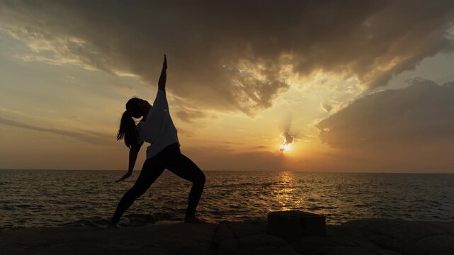 Woman practicing warrior yoga pose in silhouette at ocean sunset. Peaceful outdoor yoga scene by the sea, ideal for wellness, mindfulness, fitness, meditation and healthy lifestyle content.