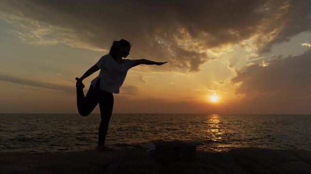 Woman practicing dancer pose yoga at sunset by the ocean, forming a graceful silhouette against a dramatic golden sky. Wellness, mindfulness and balance concept with tranquil coastal atmosphere and co