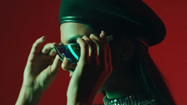 Stylish young woman wearing a beret and holding sunglasses against a red background with green cinematic lighting, ideal for fashion advertising and creative storytelling.