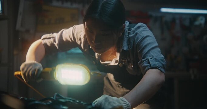 Dressed in Work Overalls and Protective Gloves, Young Female Leans Over Open Hood, Using Handheld Lamp to Illuminate Hard to Reach Areas of Engine. Expert Observation and Identifying Potential Faults.