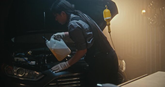Young Asian Female Mechanic in Work Overalls Carefully Pours Washer Fluid in Car Under Open Hood. In Garage, Girl Focuses on Precise Maintenance, Ensuring Optimal Vehicle Performance and Reliability.