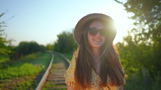 Portrait of happy woman in yellow dress smiling on railroad track. Happy girl with hat walks outdoors in summer nature. Woman faces camera with smile. Sun light shines on happy female traveler.