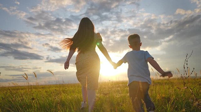 Mother and son running in green field at sunset holding hands. Happy family runs through grass during summer evening. Mother and son enjoy nature outdoors. Family goes forward in sunset field.