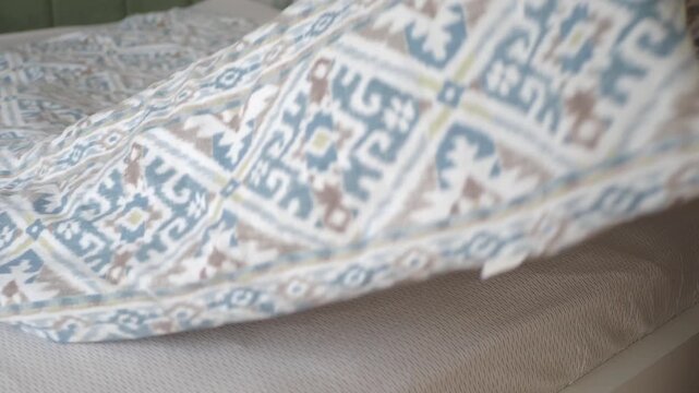 Person spreading patterned duvet cover on mattress in bright bedroom to make bed in morning