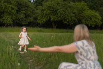 Fototapeta premium A young Caucasian girl in a white dress runs towards a woman with blonde hair in a patterned dress. They are in a green field with trees in the background.