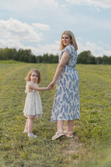 Fototapeta premium A young Caucasian woman and a small girl hold hands in a green field. The woman has long blonde hair and wears a patterned dress. The girl has short blonde hair and a white dress.