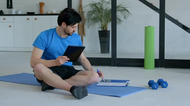 Sporty fit man sitting on exercise mat, networking online with digital tablet and planning fitness workout while preparing for sports training in domestic room.