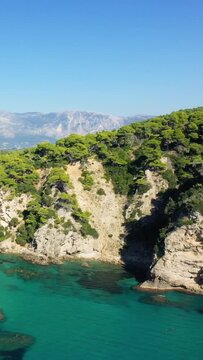 Aerial view of the rocky coastline and crystal-clear turquoise waters at Alonaki Fanariou beach, surrounded by lush green forest and distant mountains under a clear blue sky.