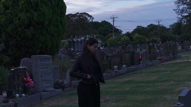 A woman stands contemplatively in a Melbourne cemetery as the sun sets, casting a soft glow over gravestones. The jib angle adds depth to the solemn scene.