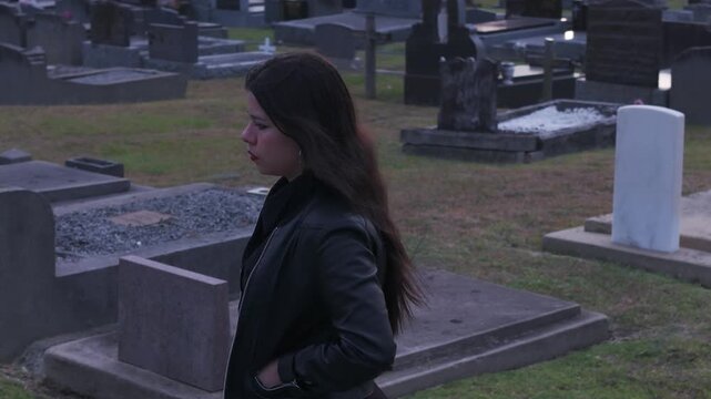 A young woman in a leather jacket stands somberly in a graveyard at sunset in Melbourne, Australia. The setting sun casts a melancholic glow over the tombstones and grassy area.
