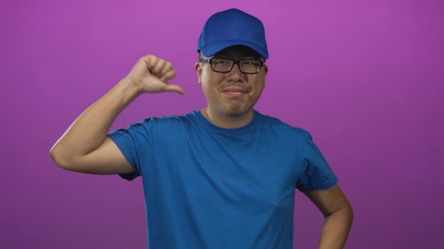 Man smiling, wearing blue cap and t shirt, showing open hands and a thumb pointing to self in a purple studio; friendly greeting.