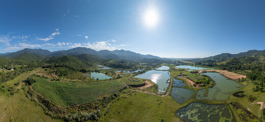 Aerial panoramic view of artificial fish ponds and wetlands surrounded by green hills and mountains in Santa Catarina, Brazil under a clear blue sky.