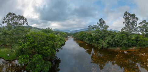 Aerial panoramic view of a calm river flowing through dense green forest with mountains in the background under a dramatic cloudy sky in Santa Catarina, Brazil.