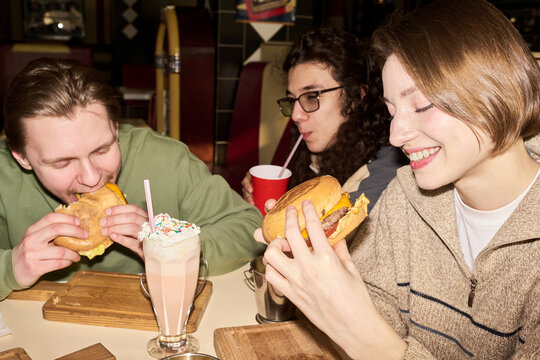 Three gen Z friends eating burgers and drinking milkshake at restaurant table, smiling and enjoying meal together