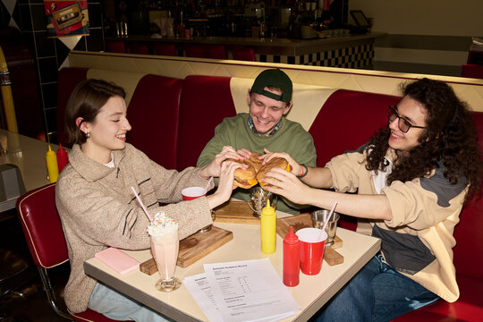 Three young friends sitting in retro diner booth sharing burgers and milkshakes, smiling and enjoying meal together, food and drinks on table
