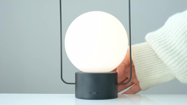 A woman's hand turns on and off round lamp standing on a table.