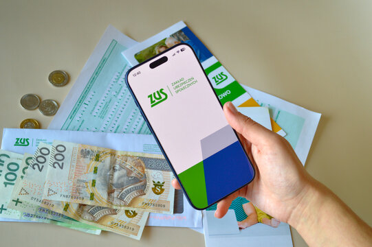 A hand holds a modern smartphone displaying the Polish ZUS social insurance logo above a table covered with zloty banknotes and official government documents for financial planning.