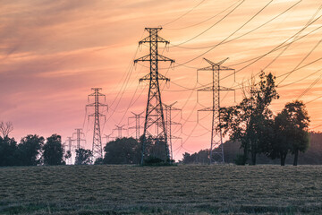 Linie wysokiego napięcia i maszty energetyczne na tle malowniczego zachodu słońca. Infrastruktura energetyczna na polu w ciepłym, pomarańczowym świetle wieczornym. © Henryk Niestrój