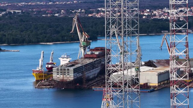 Aerial view of a shipyard with ships docked in the calm, deep blue water, with cranes standing tall against the backdrop of the coastal city, Pula, Istria County, Croatia.