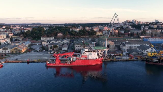 Aerial view of a massive red ship docked at the shipyard, reflecting in the still, dark water, contrasting with the urban landscape, Pula, Croatia.