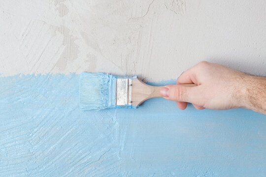 Young adult man hand holding brush and applying blue waterproofing coating on concrete wall or floor before lying tiles at bathroom. Closeup. Repair work of home. Front view.