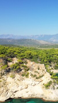 Aerial view of rugged white cliffs and lush pine forest by the turquoise sea at Alonaki Fanariou beach, Greece, with mountains in the background under a clear blue sky.