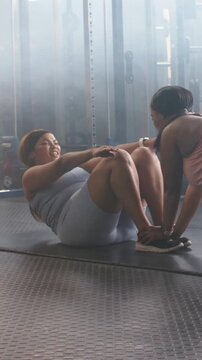 Vertical video: Doing assisted situps, woman in gray top on mat toning core, partner bracing feet