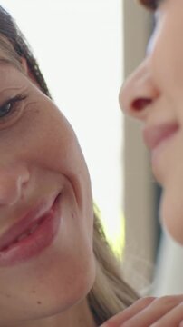 Vertical video: Daughter kissing mother's cheek at home, making her laugh with hoop earring, window