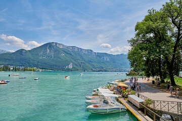 Naklejka premium Outdoor, lake and mountains with boats at jetty for tourism, sky and space at vacation resort. Promenade, hills and boardwalk with people, ship and summer with clouds, holiday or water in France