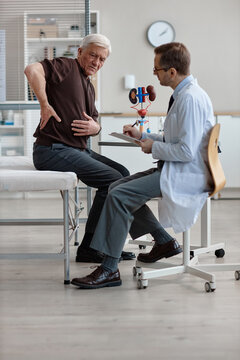 Senior Caucasian man showing lower back pain while sitting on examination table, middle aged Caucasian male urologist in coat consulting patient in medical office with kidney model