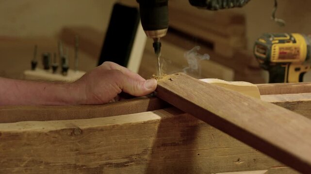 Woodworker drilling pilot hole in hardwood plank with cordless power drill and smoking wood friction