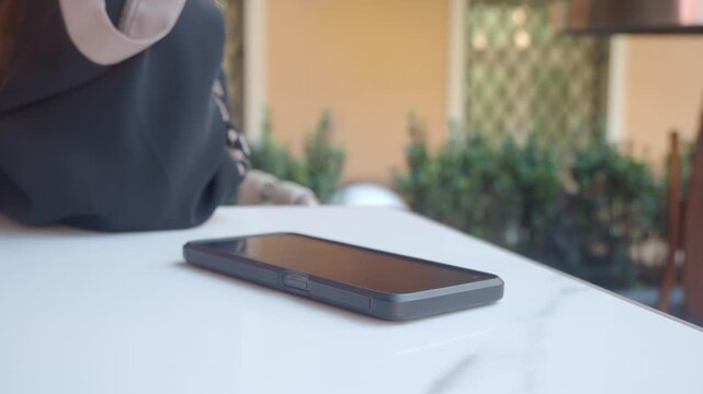 Unrecognizable woman wearing black hijab and floral patterned dress picks up smartphone from marble table