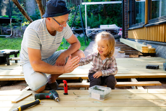 Joyful father and little child building a wooden patio deck together. Authentic family DIY home improvement project, sunny outdoor leisure.