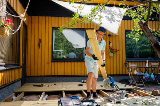Man building wooden deck in backyard. Authentic DIY home improvement project during summer. Outdoor house renovation and construction.
