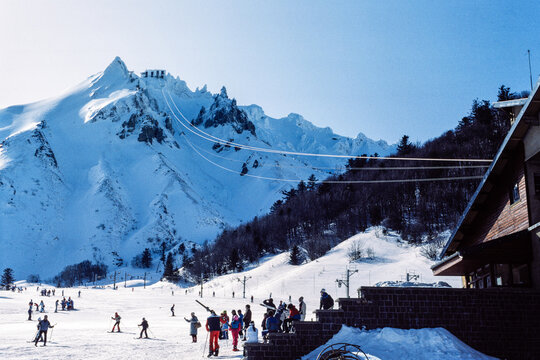 Le Mont-Dore, Puy-de-D&ocirc;me, France, February 1984: skiers on snowy slopes near chalet with cable lines leading to alpine peak in a vintage French winter resort scene