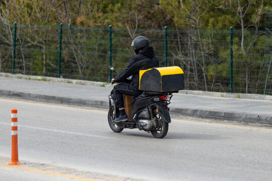 A delivery courier in full black gear and a matte helmet rides a black scooter along an urban road.