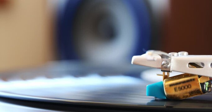 Turntable stylus on vinyl record with blurry, out of focus speaker in the background. Close up macro shot, side view, real time video, no people