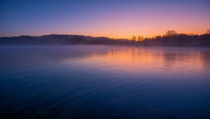 Obraz premium Reflecting calm lake surface mirroring sunrise colors at rural lakeshore, with mist and ripples