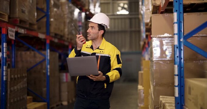 Caucasian male warehouse employee holding laptop and speaking on walkie talkie while checking stock levels of stacked cardboard boxes inside industrial logistic inventory facility