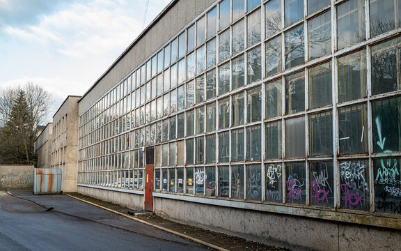 Exterior view of an old industrial building with a long glass window facade covered in graffiti. Weathered structure with repetitive grid windows and urban street environment.