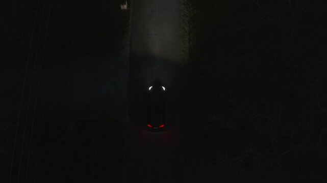 Bird's eye drone view of a single car driving through a dark, quiet residential neighborhood at night. Headlights illuminate the empty street below