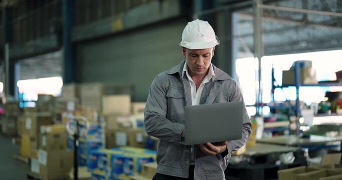 caucasian male warehouse operator using laptop to check inventory tracking system while standing near stacked boxes inside large distribution center with organized packages around