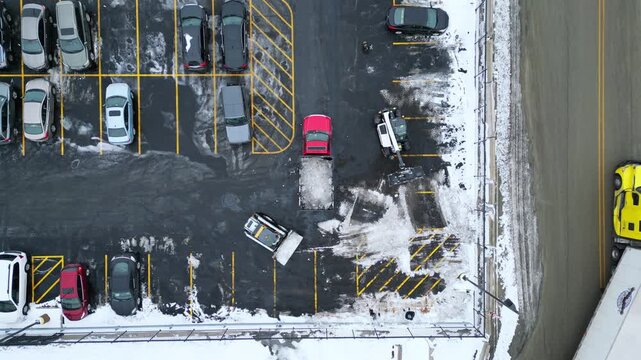 A drone captures heavy machinery and a red pickup truck clearing snow and ice from a commercial parking lot with several parked cars.