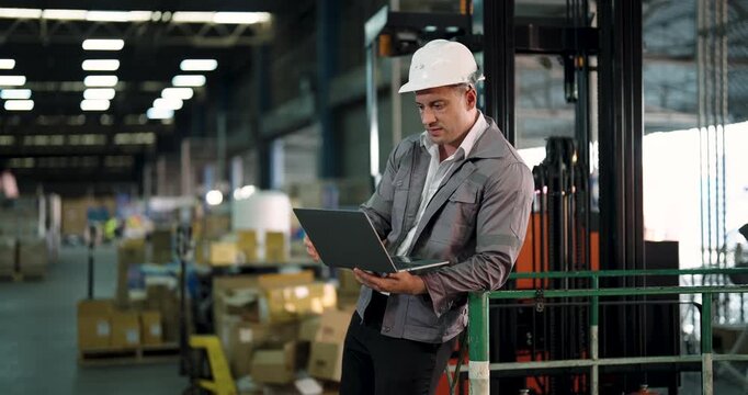 caucasian male logistics worker reviewing stock records on laptop while standing next to forklift inside industrial warehouse with cardboard boxes and stacked cargo in background