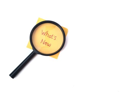 Magnifying glass over yellow sticky note with What's New text on white background. Represents discovery, business updates, market research, innovation, and staying informed